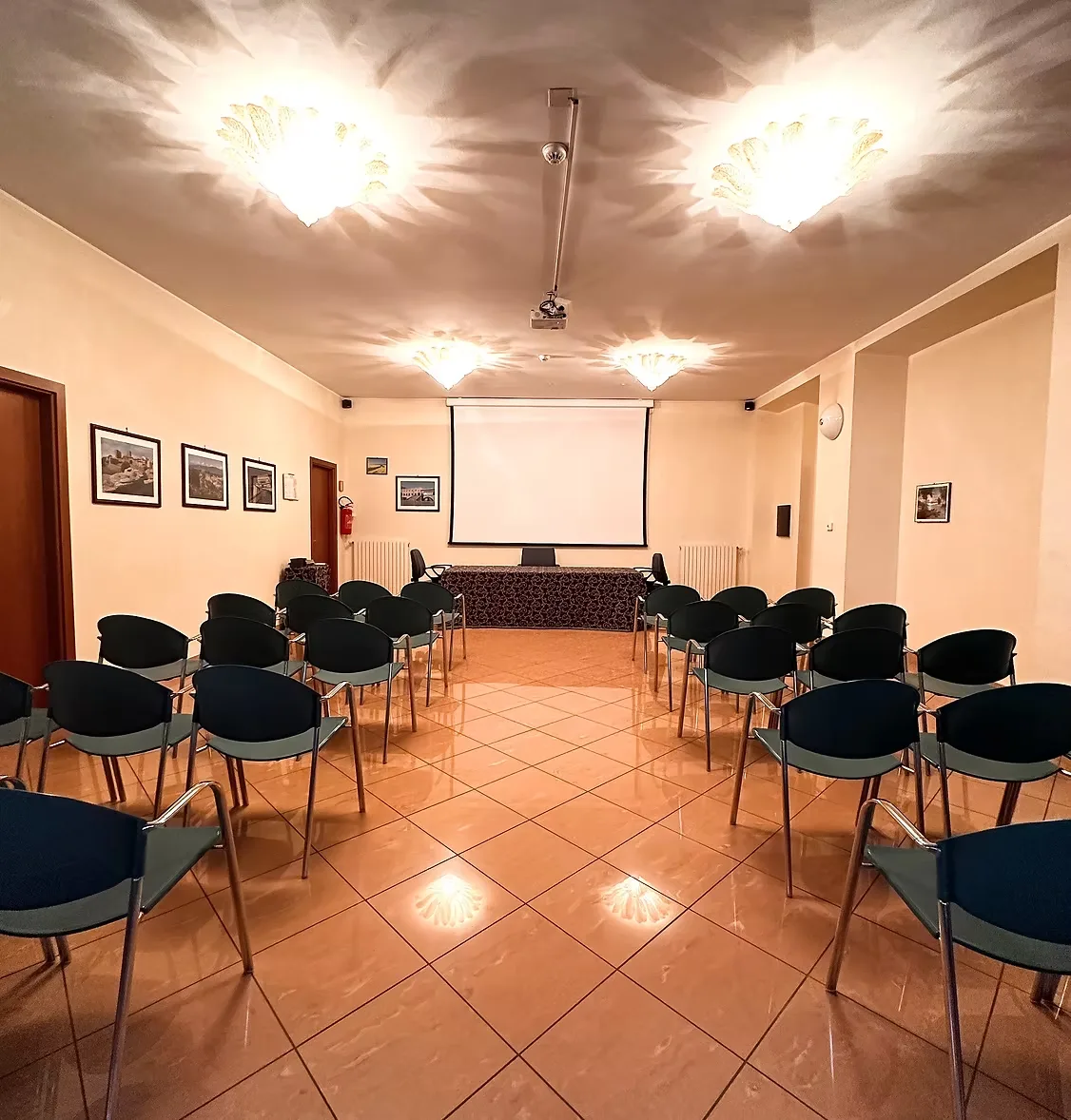 Sala conferenze con sedie a platea, schermo per proiezioni e tavolo relatori illuminato dai lampadari in vetro